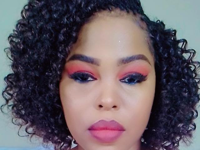 Sadeh69 has curly black hair, pink eyeshadow, blue eyeliner, and pink lipstick. Her dark brown skin is smooth, and she has a serious expression on her face.