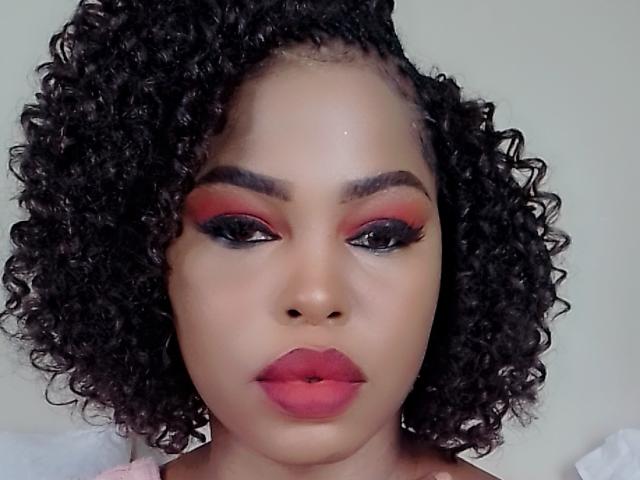 Sadeh69 has curly black hair, red and black eyeshadow, long black eyelashes, and pink lipstick. She's looking directly at the camera with a neutral expression.