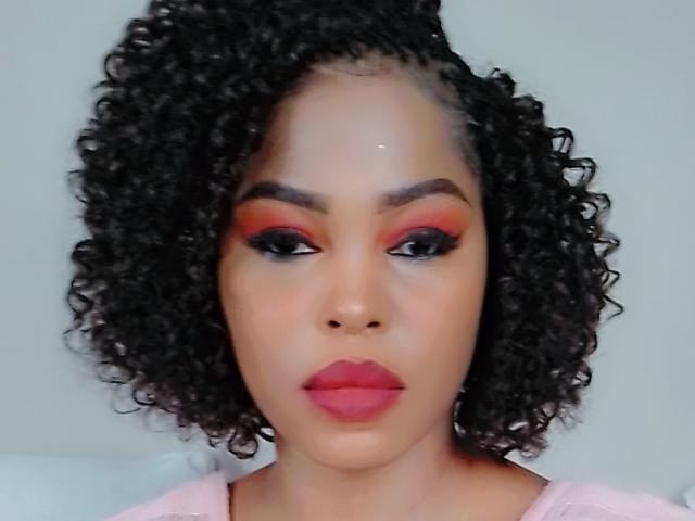 Sadeh69 has curly black hair, red eyeshadow, black eyeliner, and pink lipstick. Her brown skin is smooth, and she's wearing a pink shirt. Her ass is not visible.