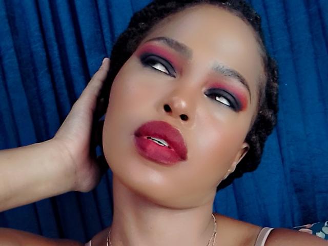 Sadeh69 has dark eyes with bold red and pink eyeshadow, full red lips, and a hand on her head. Her skin is smooth, and she's wearing a gold necklace.