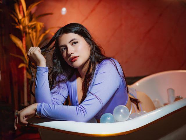MiaLeiya with long dark hair, wearing a purple top, holding her hair, looking at the camera, sitting in a bath with a sultry expression, showing her tits.
