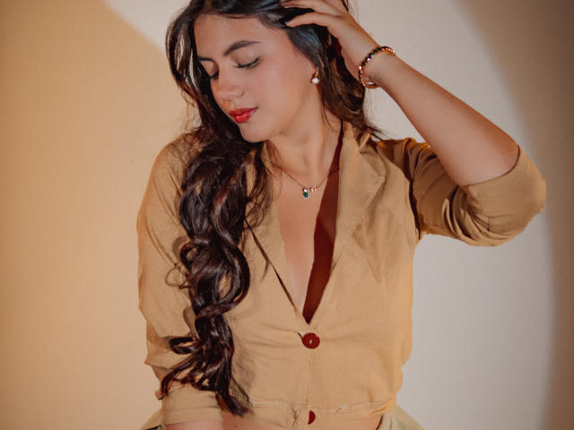 MiaLeiya with long, wavy dark hair, tan shirt, red button, and necklace, touching her hair, wearing red lipstick, and a bracelet, looking down with a subtle smile.