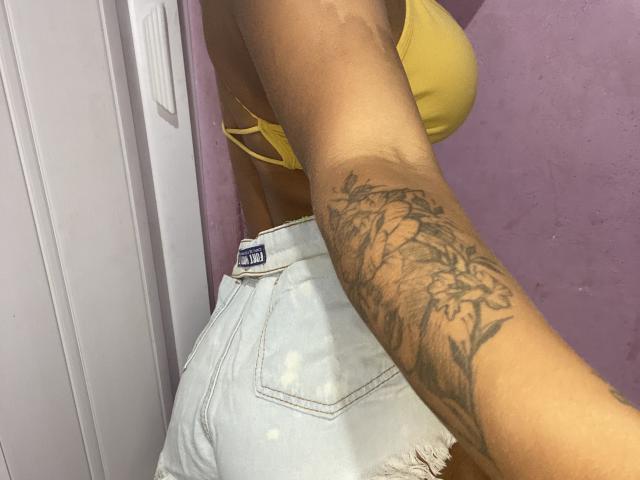 VanessaNataliia with tan skin, yellow bra, and ripped denim shorts, showcasing a detailed floral tattoo on her right arm, taken in a vertical selfie.