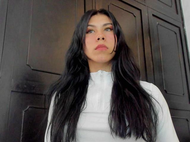 SophiePaytton with long black hair, white high-neck top, and pink makeup, looking up with a neutral expression, black wooden doors in the background.