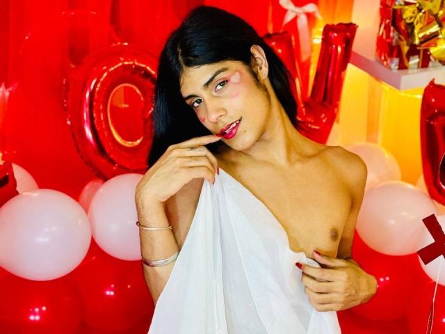 ValeryTranss with dark hair, red face paint, and red lipstick, holding a white sheet to cover their small tits, standing in front of red balloons and metallic letters.
