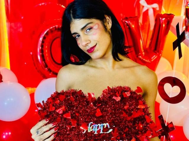 ValeryTranss with black hair, red makeup, and red tinsel-covered boobs, holding tinsel, smiling, in a red-themed setting with balloons and metallic letters.