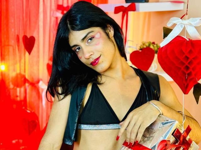 ValeryTranss with black hair, red lipstick, and pink heart makeup, wearing a black bikini top and leather vest, holding a gift with red ribbon, looking at camera.