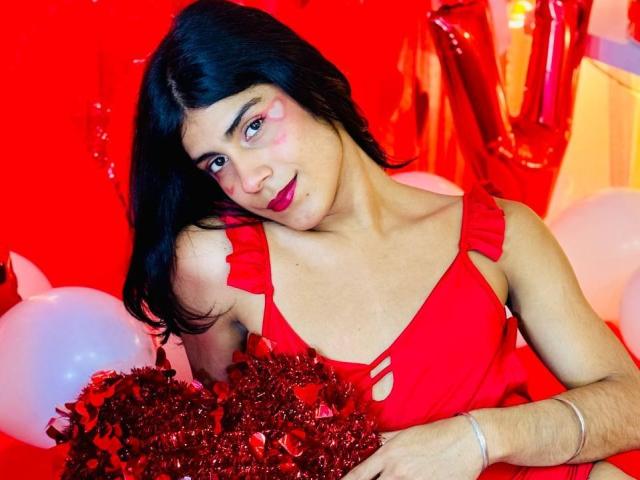 ValeryTranss with dark hair, red dress, and red lipstick, holding red tinsel, has small boobs, pink eye makeup, and a slight smile, in a red-themed setting.