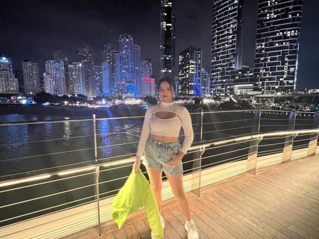 NovaDixon with big ass, standing in neon city, wearing white crop top, high-waisted denim shorts, white sneakers, holding green jacket, nighttime, city skyline, water reflection, metal railing.