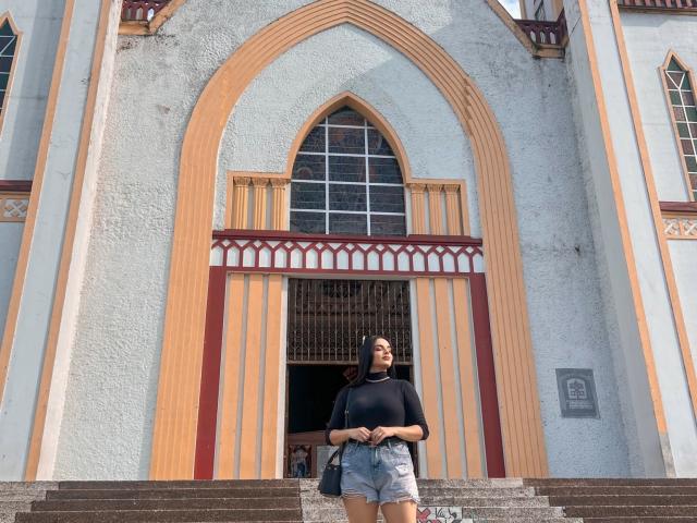 NovaDixon stands in front of a Gothic-style building with an arched window, wearing a black crop top, denim shorts, and a cap, with a bag on her shoulder.