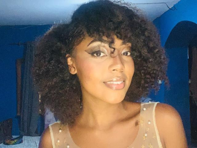 Moonisha tem cabelos cacheados e castanho-escuros, pele marrom-clara e está sorrindo com a língua ligeiramente para fora. Ela está vestindo uma blusa transparente e decorada, com um delineador marcante..
