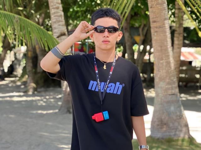 CockOfAsian, wearing black t-shirt, black sunglasses, short black hair, wristband, necklace, red-blue 3D glasses, standing, palm trees, sunny, raising right hand, looking forward, casual.