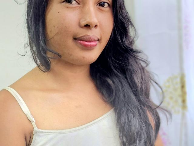 MillieJollie has medium brown skin, dark wavy hair, and is wearing a white tank top with thin straps, showing her small, cute tits and a subtle smile.