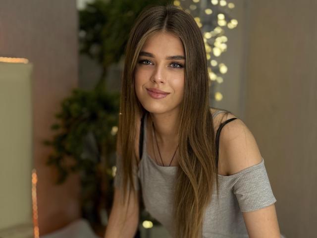 MelissaCrayzy has long brown hair, blue eyes, and a cute smile. She's wearing a grey off-shoulder top, showing black straps, and leaning forward with a subtle, confident look.