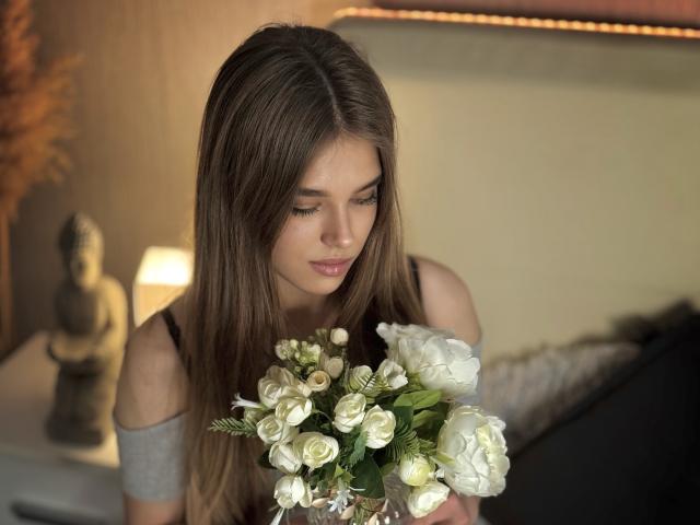 MelissaCrayzy with long brown hair, looking down at a bouquet of white roses and greenery, wearing a black top with off-shoulder sleeves, in a softly lit room.