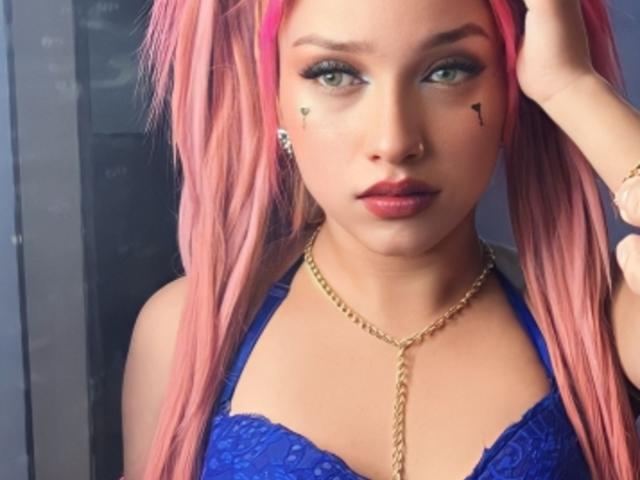 Aliceandmike69, avec des cheveux roses, un soutien-gorge en dentelle bleu, un collier en chaîne dorée, et des tatouages de cœurs noirs sur les joues, regardant l'appareil photo avec une main dans les cheveux. Yeux verts, rouge à lèvres rose..