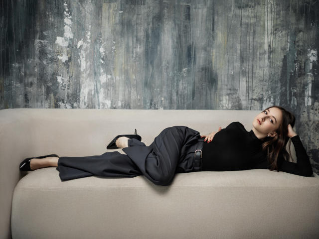 LilieFlower is laying on a beige couch, wearing a black turtleneck and gray pants, with black heels, her ass slightly raised, her hand on her head.