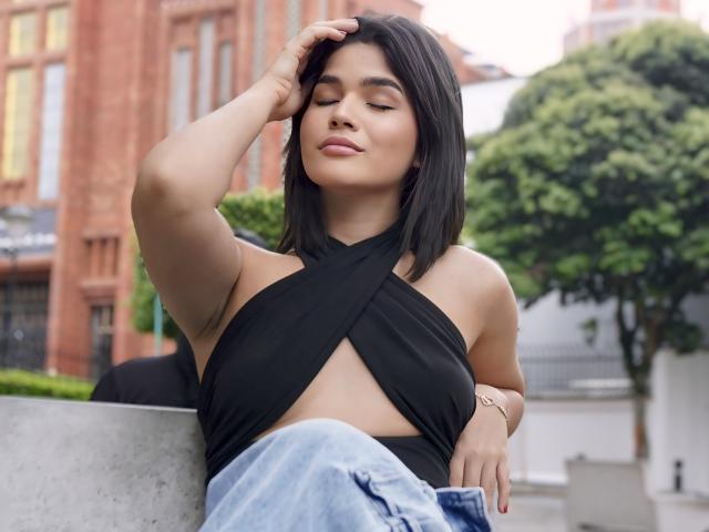 ScarlletZhen with short black hair, closed eyes, and a slight smile, wearing a black criss-cross top revealing her tits, and blue jeans, sitting with one arm on her head.