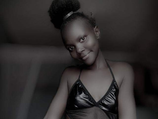 SweetSexyBabe with dark skin, curly top bun, wearing a shiny black halter top, smiling softly, showing small tits, blurred background, dark gray tone, medium shot.