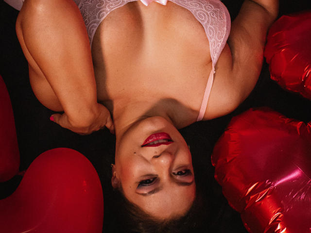 MiaCallie with red lips, white lace bra, and red heart balloons, lying on her back, arms above her head, showing her tits, surrounded by shiny red balloons.