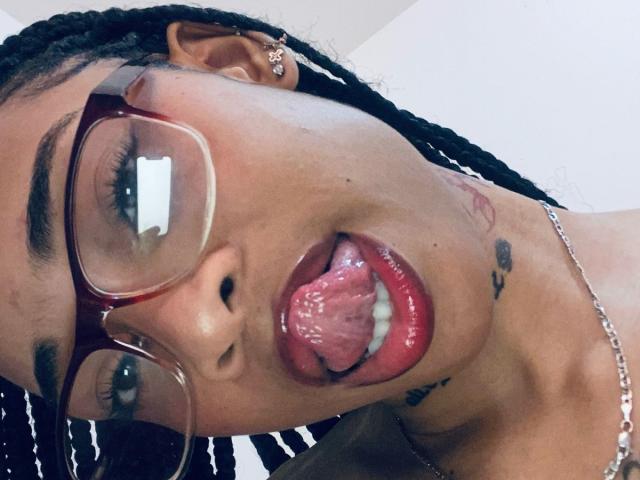 ElaHott's face with her tongue out, red lipstick, and glasses, showing a tattoo near her jaw. She has braids and a silver earring. Her tongue is wet and shiny.