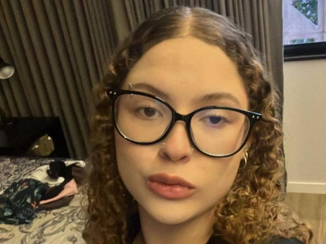 JuliaMartinsV has curly brown hair, black glasses, and a nose piercing. She's pouting with pink lipstick. Her ass isn't visible. Bedroom with clothes on bed.