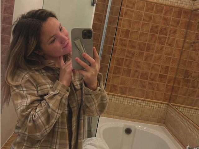 JudyGrace with pouty pink lips, holding phone, wearing plaid shirt, standing in bathroom, reflection in mirror, brown tiled walls, white bathtub, pink nail polish, blonde hair.