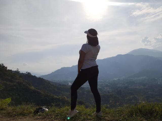 BritanyHotLady, wearing a white cap and white t-shirt, stands with her back to the camera, black pants, and white sneakers, on a grassy mountain overlook with sun overhead.