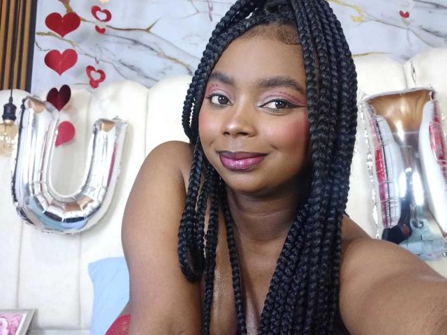 SamyLorenss has dark skin, long black braids, pink eyeshadow, purple lipstick, and a smiling face with her bare shoulders visible. She's in front of shiny "U" balloons.