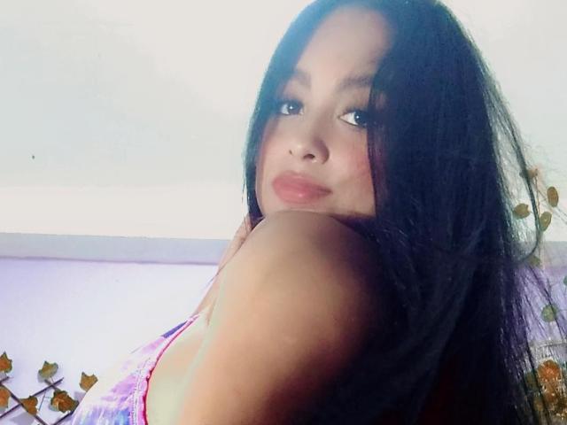 AliceBoltonX met lang zwart haar, lichtbruine huid en roze lippenstift, kijkt over haar schouder, waarbij haar blote schouder zichtbaar is, en draagt een kleurrijke top, met een subtiele glimlach..