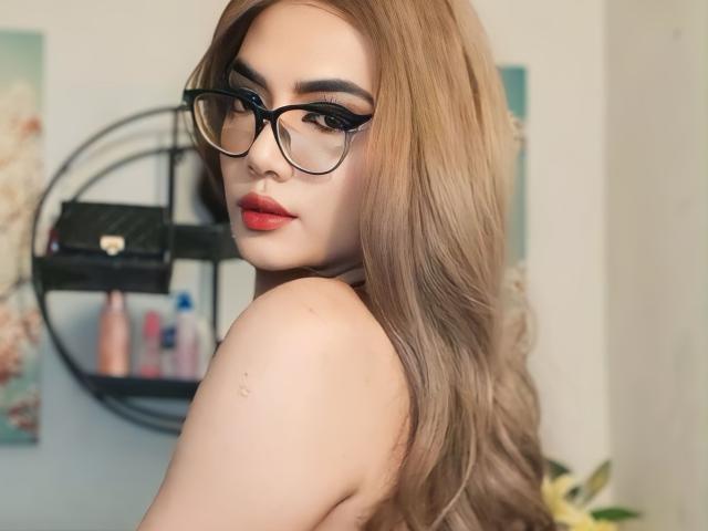 TransKathie with long brown hair, red lipstick, and black glasses, looking over shoulder, showing bare shoulders, with a subtle nose piercing. Background includes shelves with beauty products.