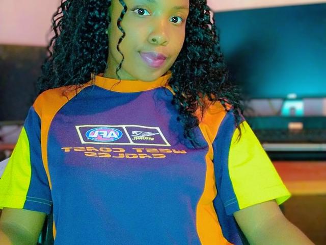 TiakoI met krullend zwart haar, gekleed in een blauw en oranje voetbalshirt met gele mouwen en het logo "Terra Tech Soccer Club", staat er met een speelse, fronsende uitdrukking..
