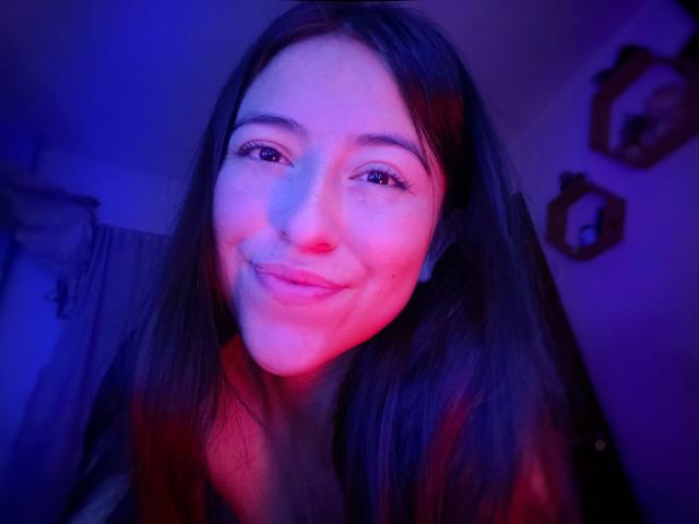 KatiaRogers with long brown hair, blue and pink lit face, slight smile, wearing a dark top, blue and red light reflecting on her skin.
