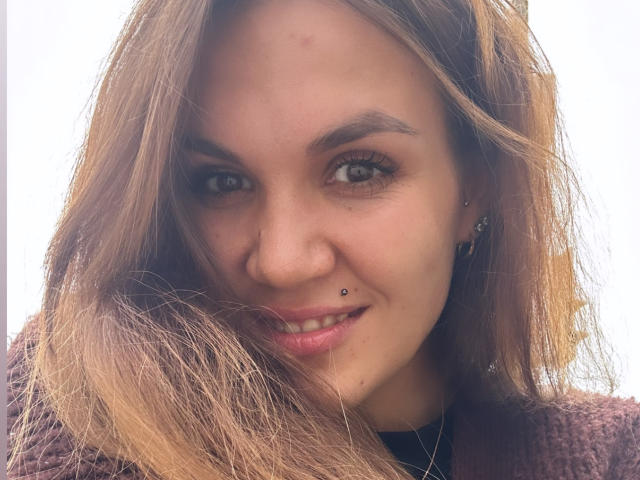 SashaWild, con cabello castaño claro, un piercing en la nariz (un pequeño aro negro) y aretes, sonriendo a la cámara, vestida con un suéter marrón texturizado..