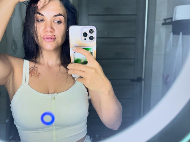 SashaWild, met donker haar, roze lippen en een tatoeage op haar rechter schouder, maakt een foto van zichzelf in een spiegel, gekleed in een witte tanktop, waarbij haar borsten zichtbaar zijn, en ze houdt een telefoon vast..