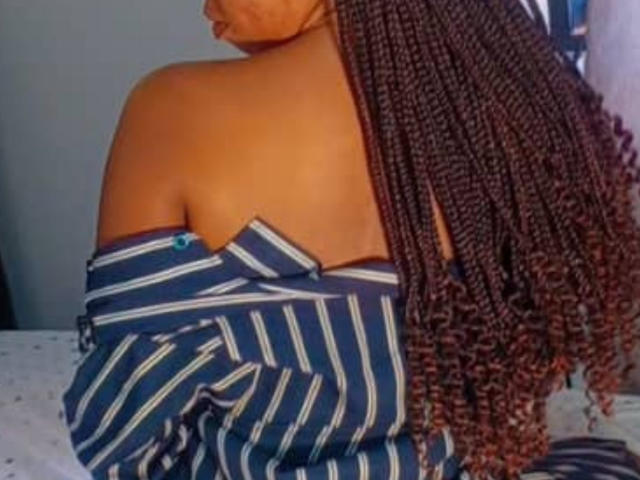 BellaMiaX with brown skin, long braided hair, bare shoulder, striped shirt off, showing her tits, sitting on white bed, soft lighting, casual, relaxed, intimate.