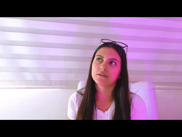 MiaLeiya with long brown hair, white shirt, and black eyeglasses on head, looking to the right with pink and purple light on her face. She's sitting on a white chair.