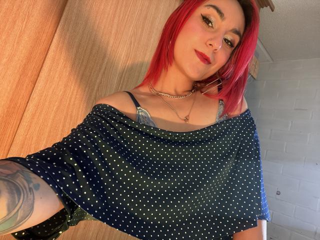 AmyStreetwise69 mit roten Haaren, trägt ein blaues, gepunktetes Oberteil mit einem Schulterauschnitt, das ihre kleinen Brüste zeigt und hat ein Tattoo am Arm. Sie lächelt in die Kamera..