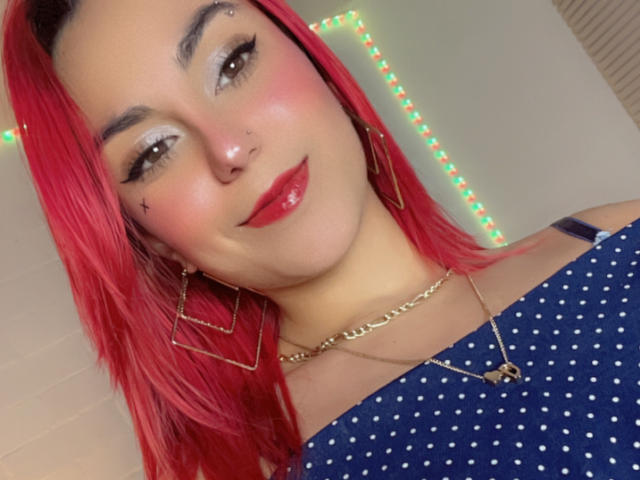 AmyStreetwise69 with red hair, nose piercing, and star tattoo, wearing a blue polka dot top, gold necklace, and earrings, smiling with red lipstick, and subtle eye makeup.