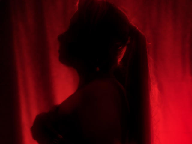 Silhouetted side profile of AnastasiaTaylor with long hair in a ponytail, covering her tits with one hand, bathed in red light. Her ass is slightly visible.
