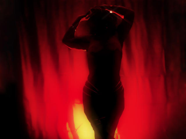 Silhouetted AnastasiaTaylor with arms raised, wearing a tight dress, standing against a bright red and yellow light, showing her ass and curves in a dark, smoky environment.