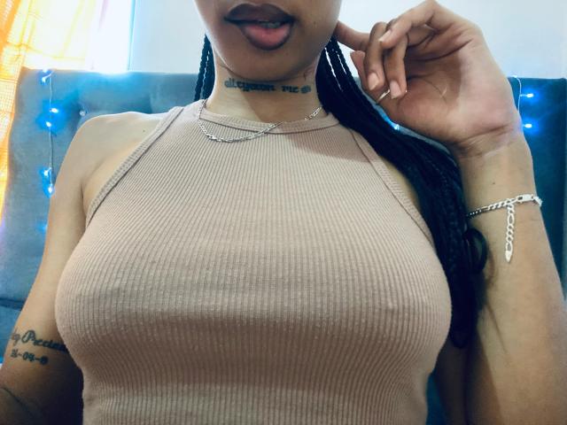 ElaHott with large, ribbed, tan top, showing her big, round tits, black braids, tongue out, silver necklace, and tattooed arm, holding her hair. Blue fairy lights.