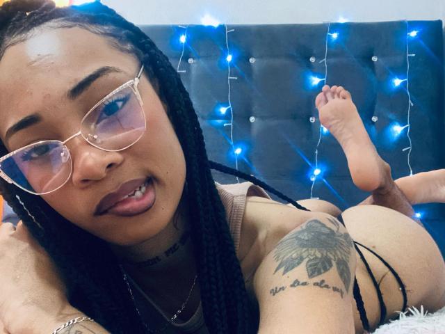 ElaHott with braids, glasses, and tongue out, lying on bed, showing her ass, tattooed arm, and foot, wearing a black thong, with blue fairy lights in the background.