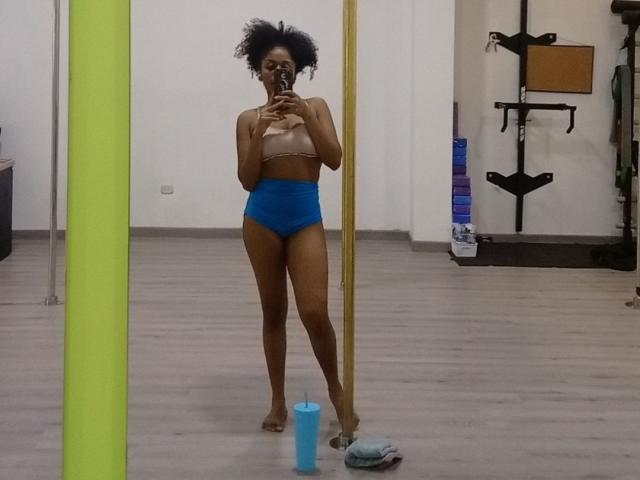 MeganWilliams with dark skin and afro, wearing a tan sports bra and blue high-waisted panties, taking a mirror selfie in a gym. A blue water bottle is on the floor.