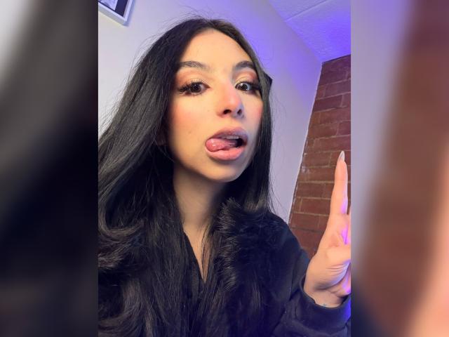 CamilleRios with long black hair, tongue out, licking her lips, wearing a black shirt, raising her finger, looking at the camera. Her face is slightly flushed.