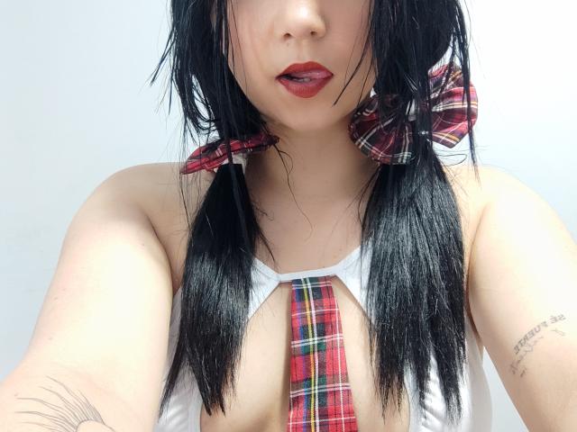 Kkatalina with black hair, red plaid bow, and white top, showing her tits, red plaid tie, and red lipstick, tattoo on right arm, white background.