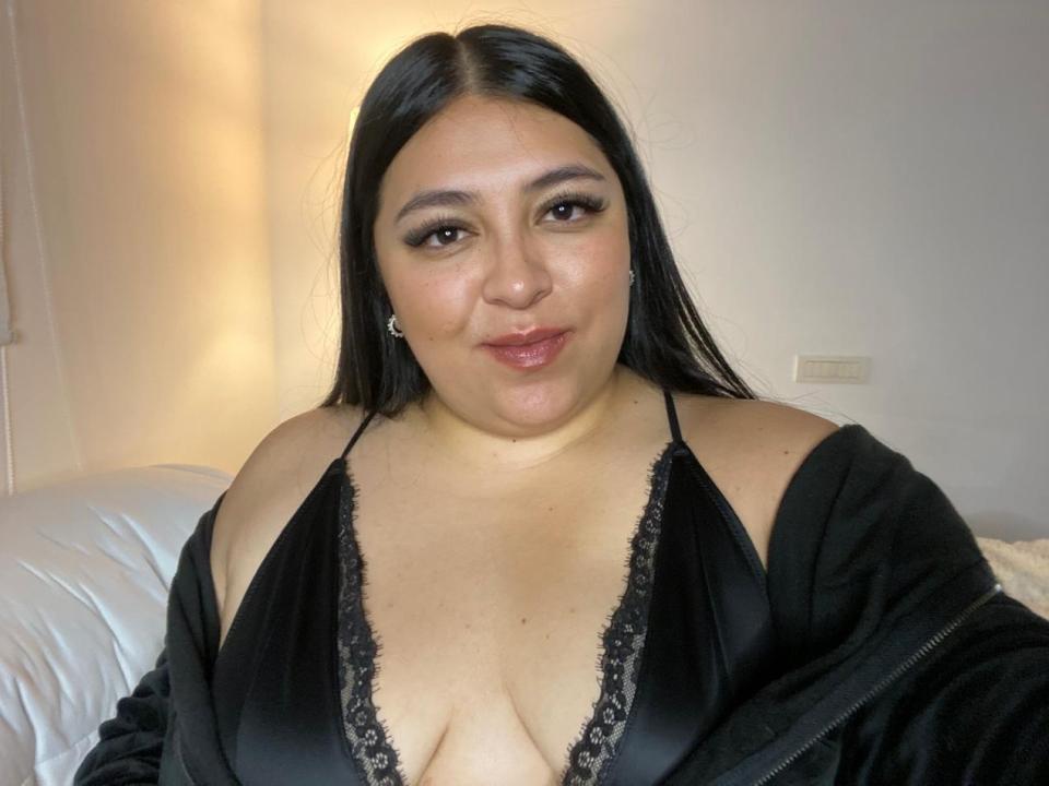 AlyFiore with long black hair, brown eyes, and full lips, wearing a black lace-trimmed deep cleavage dress, and a black jacket, showing her big tits.