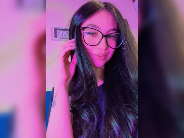 CamilleRios with long black hair, purple-pink lighting, wearing glasses, pouty expression, touching ear, black shirt, tattoo on forearm, blurred foreground, selfie-style.