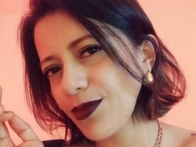 AbbyCollings a les cheveux noirs et ondulés, un rouge à lèvres foncé, des boucles d'oreilles dorées, et elle sourit légèrement, une main près de son visage, portant un collier en or sur un fond rose..