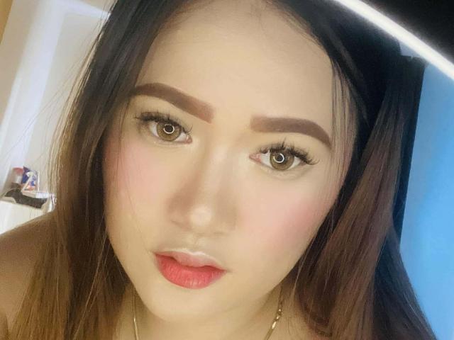 AdriannaFowler has brown eyes, thick eyebrows, long brown hair, pink blush, red lipstick, and a gold necklace. She's looking at the camera with a slightly pouty expression.